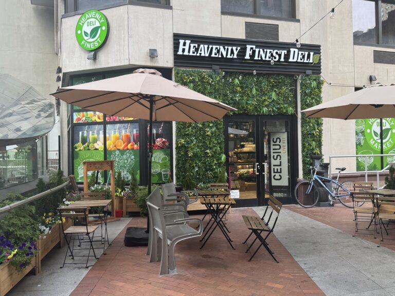 Photo shows the entrance to Heavenly Finest Deli with tables and chairs set up in a public space with a bench.