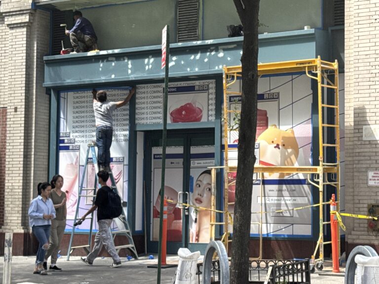 Photo shows men painting a storefront, whose windows are covered in advertisements for "SUKOSHI."