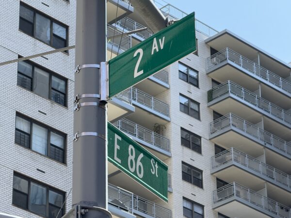 Photo shows green street signs reading 86 St and 2 Av in front of a white luxury apartment building.