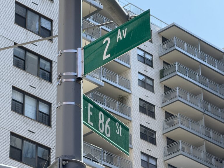 Photo shows green street signs reading 86 St and 2 Av in front of a white luxury apartment building.