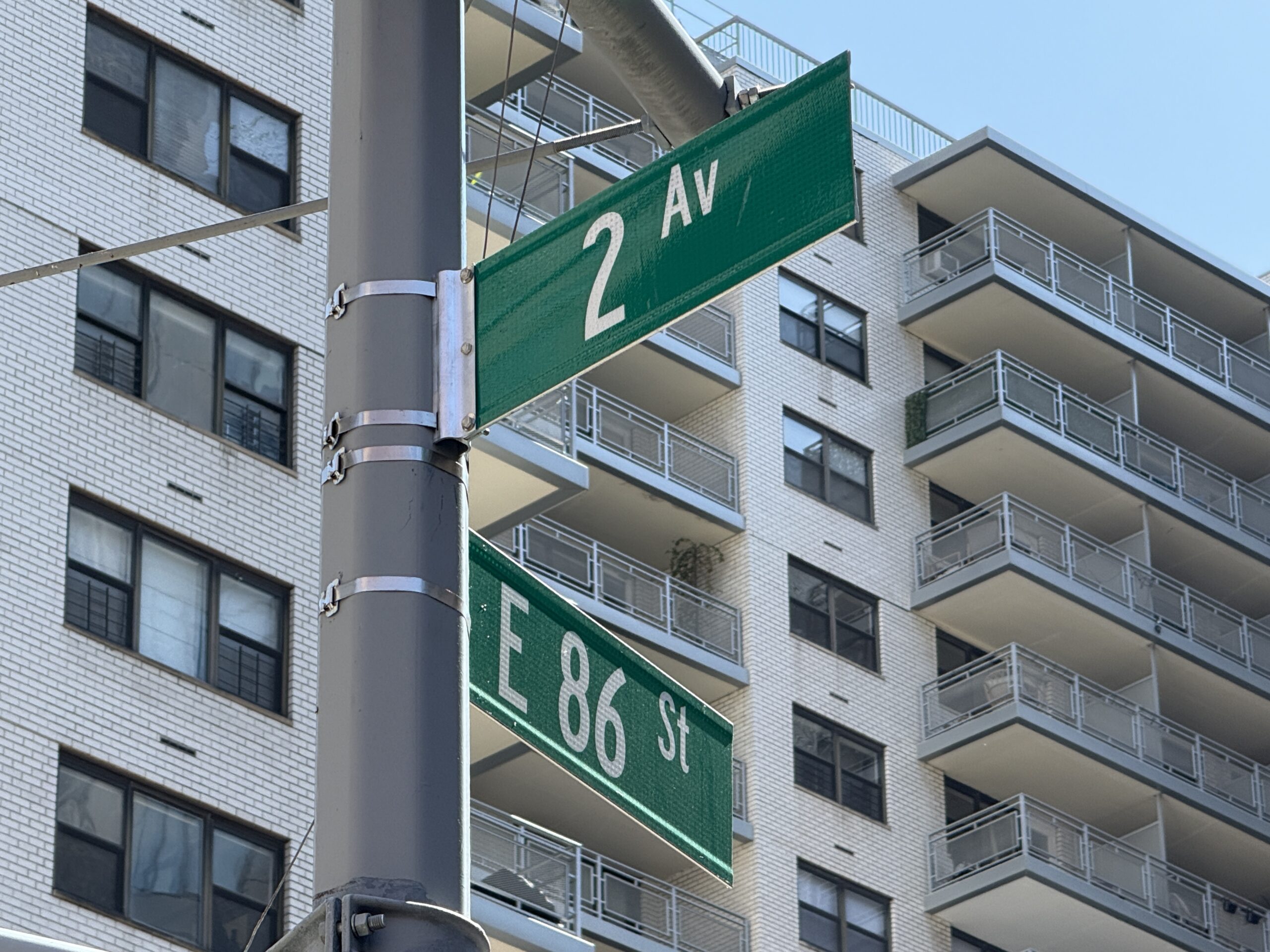 Photo shows green street signs reading 86 St and 2 Av in front of a white luxury apartment building.