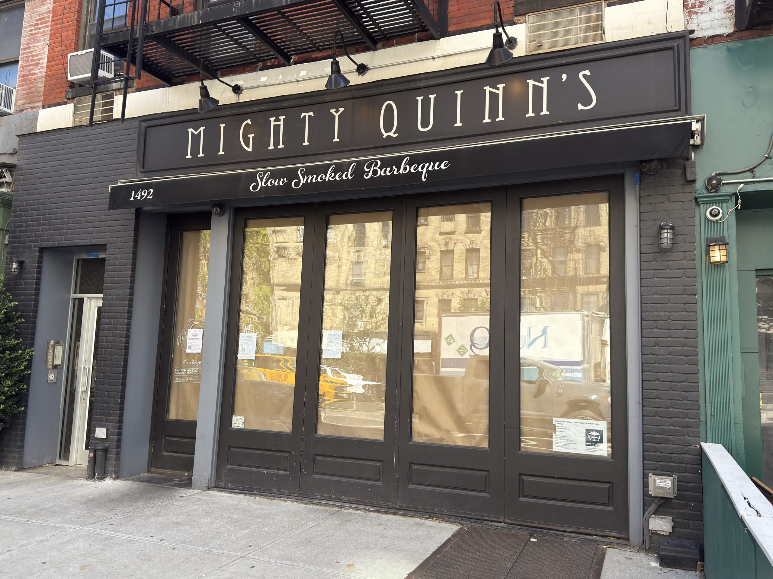 Photo shows a black storefront featuring white lettering at the top reading "Mighty Quinn's," whose windows are covered with brown paper.